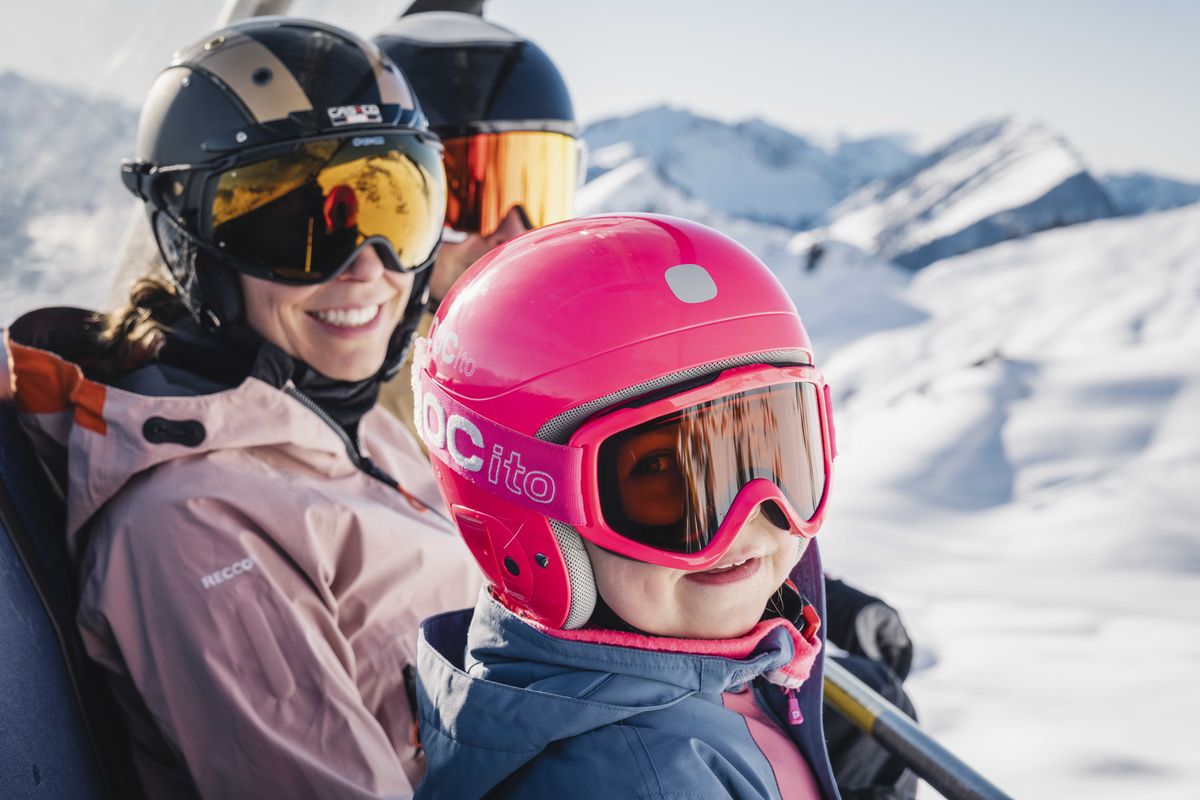 Erwachsene und Kind in Skiausrüstung mit Helm und Skibrille lachen auf der Piste, dahinter verschneite Berge